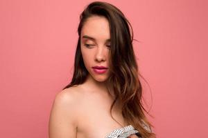 Tender stylish girl with wavy brown hair and pink lips is looking down over pink background. Portrait of charming lady is dressed in dress with bare shoulders and posing on isolated background photo