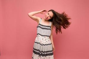 Happy excited romantic girl with wavy brown hair wearing light striped dress is dancing and having fun with flying hair over pink background photo