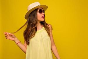foto de estudio de una joven bonita con el pelo ondulado vestido con un vestido ligero de verano y un sombrero con gafas de sol posando sobre fondo amarillo y mirando a un lado, vacaciones de verano, estado de ánimo de viaje
