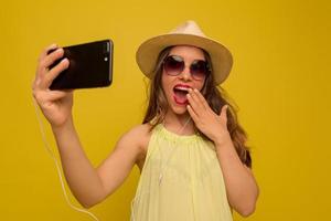 Una chica encantadora y emocionada con el pelo largo y ondulado con gafas de sol y sombrero está cubriendo los labios con la mano y haciendo selfie en el smartphone foto