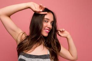 Un retrato cercano de una adorable y linda chica con cabello castaño y una sonrisa maravillosa con labios rosados toca el cabello y mira la cámara sobre un fondo rosado foto