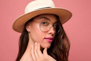 Close up tender portrait of lovable girl with great make up wearing glasses and hat is looking at camera over pink background photo