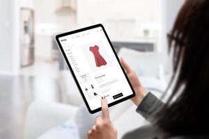 Woman buys a dress online with tablet concept. Modern e-commerce clothing website. Living room in background