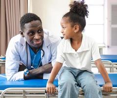 niño paciente sentado en la cama del hospital con un médico africano para recibir atención médica. niña sonriente feliz saludable después de un chequeo profesional en la clínica. practicante, pediatra revisando, examinando niño