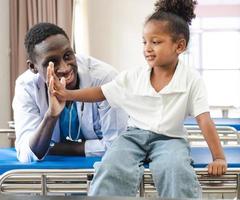 niño paciente sentado en la cama del hospital con un médico africano para recibir atención médica. niña sonriente feliz saludable después de un chequeo profesional en la clínica. practicante, pediatra revisando, examinando niño