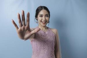 Young Asian woman wearing modern kebaya over blue isolated background doing stop sign with palm of the hand. photo