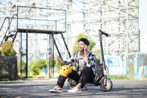 Asian man talking on the phone while sitting on electric scooter at the park street. Modern and ecological transportation concept. photo