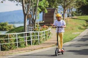 retrato de un niño asiático que usa casco disfruta divirtiéndose montando scooter eléctrico en el parque de la calle al aire libre en un día soleado. actividades deportivas saludables para niños al aire libre. foto