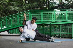 joven asiático ayudando a su esposa a hacer tablones en una alfombra de yoga en el parque. concepto de pareja de estilo de vida saludable. foto