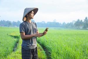 joven agricultor asiático de pie y mirando el campo de arroz mientras revisa el informe de agricultura usando un teléfono inteligente. concepto de agricultura moderna.