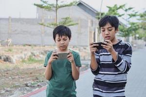 dos niños con aparatos. hermanos navegando por la red o jugando juegos en línea en teléfonos inteligentes y tabletas digitales afuera mientras están de pie. concepto moderno de comunicación y adicción a los gadgets. foto