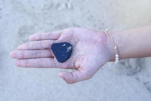 Close up of young woman's hand holding black pebble stone on beach's palm to show someone at sunrise in summer holidays, picking up pebbles on stone beach, summer vacation souvenir, selective focus photo