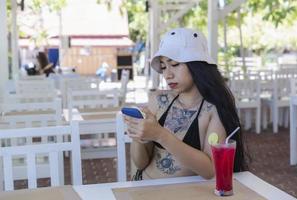 Young beautiful sexy woman in Bikini with fresh juice in restaurant using mobile phone in cafe. Pretty tattooed woman drinking tasty sweet red cocktail, amazing relaxing day on vacation in summertime photo