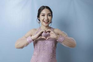 An attractive young Asian woman wearing a modern kebaya feels happy and a romantic shapes heart gesture expresses tender feelings photo