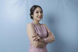 Portrait of a confident smiling girl standing with arms folded and looking at camera isolated over blue background, wearing modern kebaya dress photo