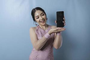 un retrato de una mujer asiática alegre que muestra espacio de copia en su teléfono inteligente sobre fondo azul, usando un kebaya moderno foto