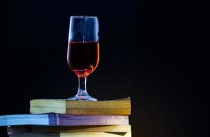 los libros antiguos se superponen en un fondo negro y con poca luz una copa de vino tinto encima. foto