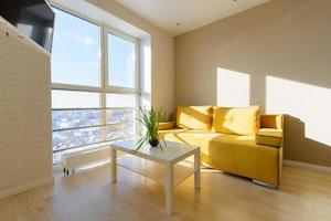 moderno y acogedor interior de apartamento, sala de estar con sofá amarillo, mesa baja blanca y tv en la pared, ventana panorámica con hermosa vista a la ciudad.