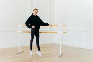 foto completa de una alegre pelirroja delgada parada de puntillas cerca de la barra de ballet, tiene ejercicios de pilates, se mantiene en forma, sostiene la mano en la cintura, usa ropa deportiva negra, posa en un espacioso salón blanco