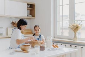 mujer morena con sonrisa le muestra a su hija pequeña cómo cocinar, da lecciones culinarias, compra diferentes productos en la tienda para cocinar, usa delantales, sonríe agradablemente, ventana con jarrón, interior de cocina foto