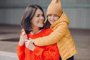 Photo of pleased attractive young mother looks positively at daughter, embraces mother, enjoy togetherness, have toothy smiles, eyes full of happiness, have good relationships. Family portrait