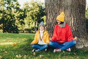 Body language concept. Carefree little kid sits near her beautiful mother, keep hands in mudra sign, keep eyes closed, try to calm down after hard work at home, admire fresh air in beautiful park photo