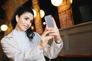 Beautiful woman with dark hair tied in pony tail making selfie while sitting in cafe using her smartphone. Well-groomed female in elegant clothes looking in screen of her telephone making photo