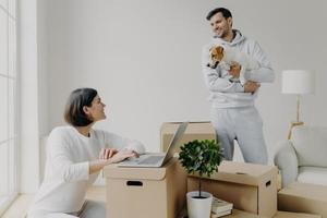 Happy family couple move into new flat for living. Wife works on laptop computer, searches information in internet, makes shopping online, man stands with dog, unpacks belongings from boxes. photo