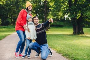 Pleasant looking dad shows with fore finger beautiful landscape to her daughter, embraces her. Family of three members have walk at park, have good relatioship, look into distance as notice something photo