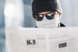 Close up shot of adult man wears sunglasses, hat and sterile medical mask, reads newspaper, finds out news about situation in world, coronavirus spread, poses outdoor against blurred background