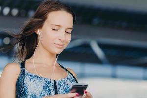 Close up portrait of attractive young woman holds smart phone, texts feedback, checks email box, listens music in player, downloads news songs, connected to wireless internet. People, leisure concept photo