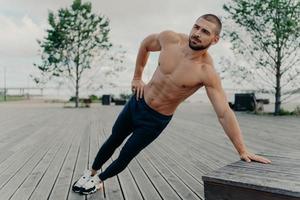 Young handsome shirtless ordinary bearded man stands in diagonal body position, makes side plank, practices endurance and stays in good physical shape. Sportsman in active wear trains outdoor photo