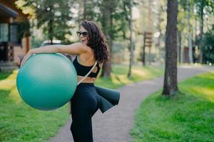 Outdoor shot of happy brunette woman carries fitball and fitness mat, wears sunglasses and sport clothes, poses in green park, being in good physical shape. Healthy lifestyle and hobby concept photo