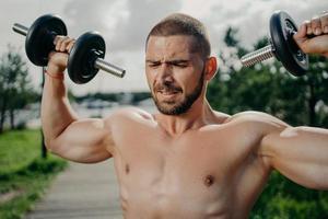 Sporty determined muscular European man exercsises with dumbbells, makes weightlifting outdoor, has naked torso, trains muscles, wants to have athletic body. Strength and motivation concept. photo