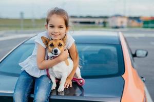 Little girl with appealing appearance, hugs her favourite pet, have journey together with parents by car, sit at trunk, pose for making photo. Children, animals, rest and transportation concept photo