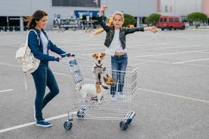 Shopping and family concept. Glad female carries trolley in which little girl stands with dogs, pose against shopping mall background, rejoice new purchases. People, happiness, day off concept photo