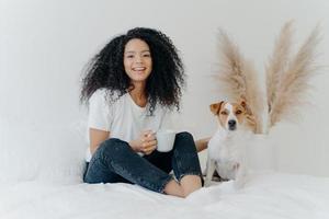 Happy carefree woman with Afro hairstyle, wears t shirt nad jeans, feels glad, holds mug of coffee, laughs sincerely, jack russell terrier poses near host. Cheerful teenage girl with favourite pet photo