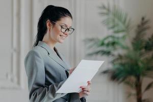 Glad female lawyer writes in corporate paper documents, wears optical glasses, formal clothes, stands in own cabinet, develops new strategy, has happy expression. People, career, employment concept photo