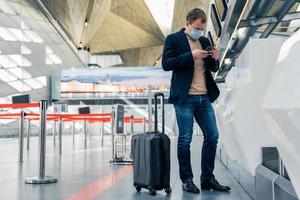 Full length shot of man passenger stands near suitcase on wheels in airport reads news online on cellular wears protective medical mask finds out about closing of countries borders because of virus photo