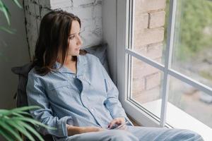 Horizontal shot of thoughtful young woman sits on windowsill, dressed in casual nightwear, uses smart phone and earphones, listens calm music while looks thoughtfully in window, enjoys home time photo