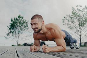 Un hombre barbudo sin camisa y con el torso desnudo se para en una pose de tablón y sonríe alegremente, demuestra resistencia, posa al aire libre, piensa en su cuerpo y su salud. chico atlético entrena afuera foto