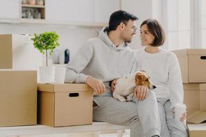 Cheerful woman and man look at each other with love, have pleasant smiles, wear casual outfit, pose with domestic animal around cardboard boxes, drink takeaway coffee, move in new cozy dwelling photo