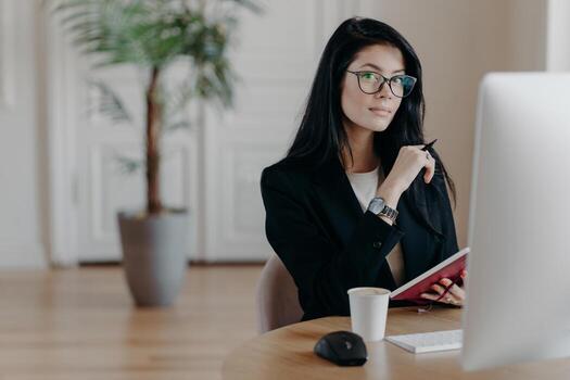 Photo of serious woman writes information in notebook, studies new online business course, poses in coworking space with computer, works on project, writes some ideas. Business and work concept