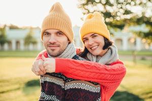 Smiling glad young female in hat and warm cotton sweater embraces her husband who stands back, enjoy spending weekends together, have walk in garden or park, admire splendid sunny autumn weather photo