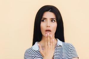 Headshot of scared brunette female covers mouth with hand, looks with fear aside, being frightened to hear unexpected news, wears casual outfit, has manicure and make up, poses over studio wall photo