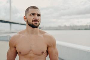 Horizontal shot of muscular man with naked torso thick beard looks somewhere into distance, relaxes outdoors, stands against river background, takes break after jogging, has athletic body shape photo