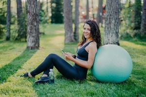 Photo of satisfied brunette young woman sits back to big fitness ball, uses mobile phone for listening music, poses on green grass in forest during sunny day, wears sports clothes. Sport concept