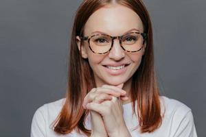 Close up shot of happy young European woman has pleasant smile, keeps hands together near chin, wears casual white clothes, isolated over grey background, being in high spirit, enjoys spare time photo