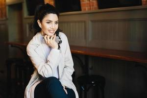 Pensive brunette female with appealing appearance looking aside while sitting in cafe enjoying favourite tracks on headphones. Relaxed woman in fashionable clothes using modern gadget indoors photo