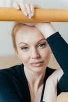 Close up shot of restful woman with ginger hair, has make up, looks at camera, has serious expression, holds hand on handrail, busy doing gymnastic exercises, has natural beauty. Ballet concept photo